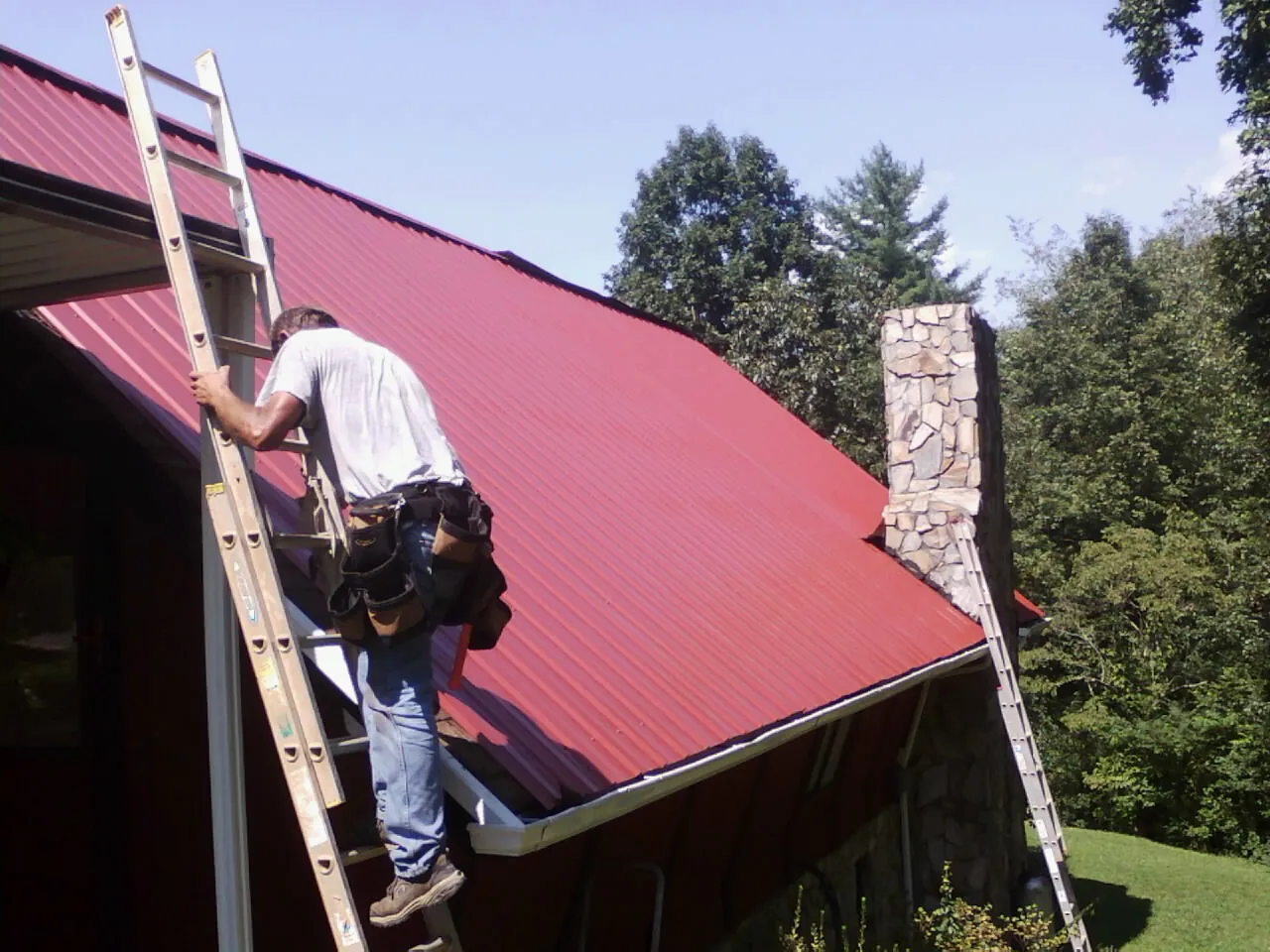 Roofer on a ladder beside a red metal roof and stone chimney — Soffit & Fascia Repair by Roofing Brothers in Columbiana